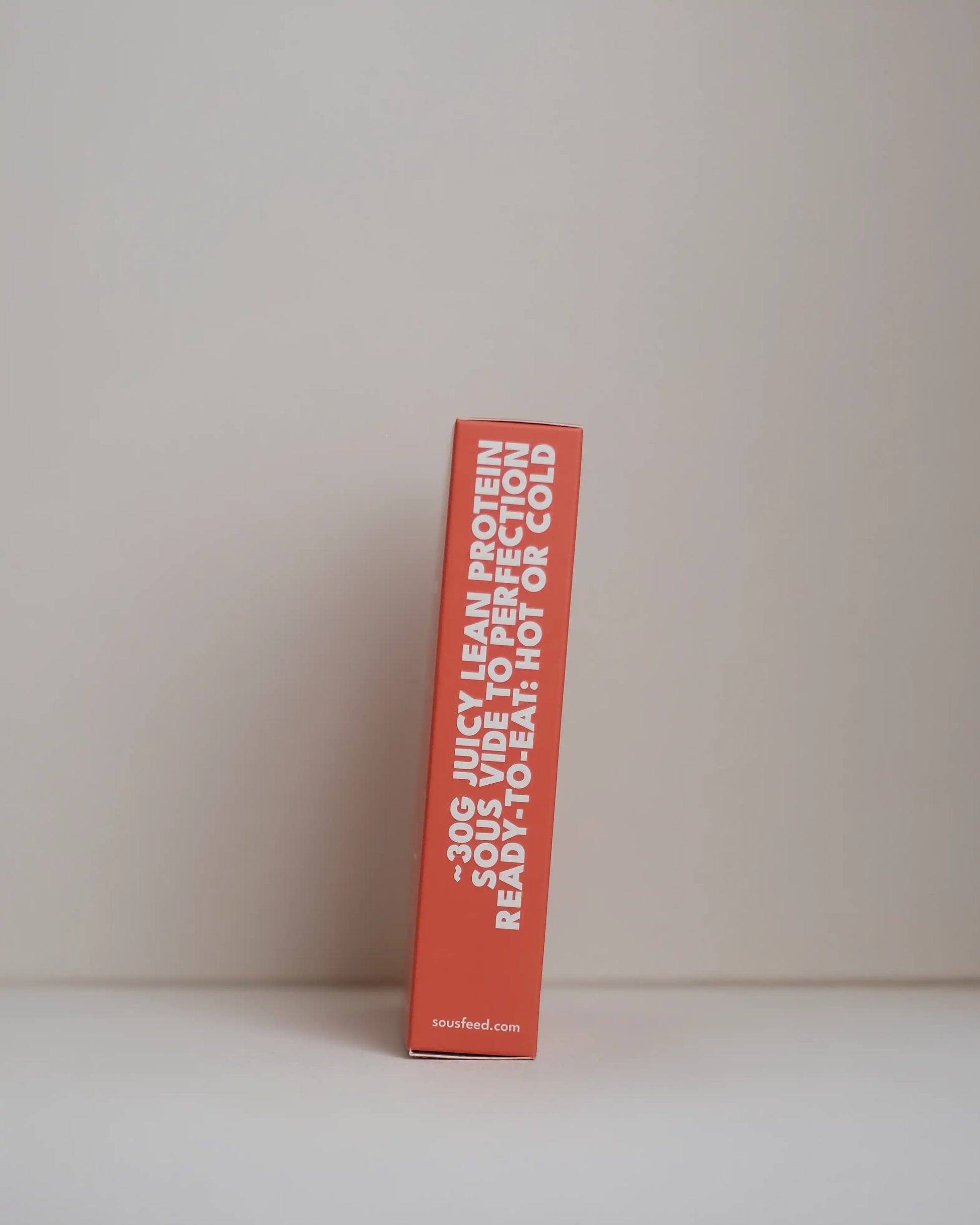 Side view of a SousFeed smoked harissa flavoured chicken breast and its red packaging on a white background