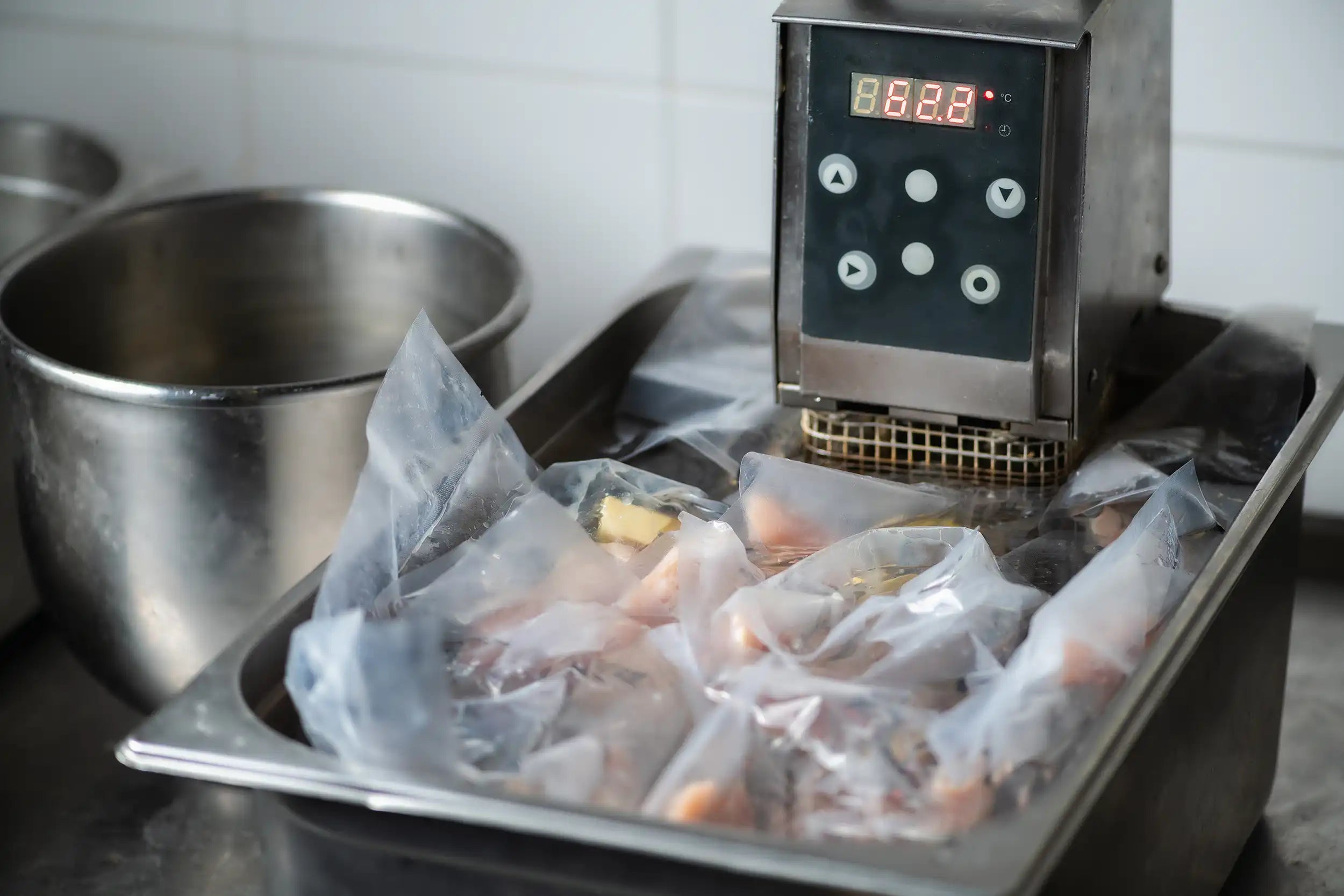 metal container with a sous vide immersion circulator and vacuumed packed chicken being cooked in the water bath.